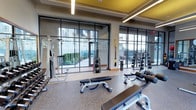 This contemporary fitness center is designed to support a full range of workout routines in a bright, motivating environment. The space is defined by its striking floor-to-ceiling glass wall system, which floods the interior with natural light and offers views of a landscaped outdoor courtyard, creating an open and energizing atmosphere. The flooring is a durable, cushioned rubberized surface in a speckled finish, well-suited for high-traffic gym use and providing comfort underfoot during exercise. Overhead, linear LED lighting is recessed into a coffered ceiling accented with bold yellow panels, adding a modern, dynamic touch to the overall design. The fitness center is outfitted with a comprehensive selection of equipment, including weight benches, adjustable dumbbells and a dumbbell rack, cable machines, a multi-function strength station, and TRX suspension training straps. The layout is thoughtfully organized to allow users to move freely between stations without congestion. Large glass doors provide direct access to the adjacent outdoor area, seamlessly connecting the indoor fitness space with the exterior environment and enhancing ventilation and natural airflow throughout the room.