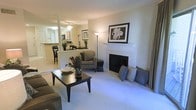 This single-floor residence offers an open-concept layout that connects the living area, dining space, and kitchen into one cohesive environment. The living room is anchored by a white-mantled fireplace, which serves as the room's focal point, flanked by decorative pillows and accented with framed wall art above. Plush wall-to-wall carpeting runs throughout the living area, contributing to a warm and comfortable ambiance. A dark wood coffee table and patterned armchair add contrast to the neutral tones of the space.

A partial wall with a structural column separates the living area from the kitchen, which is visible through a pass-through opening and features a raised breakfast bar with seating. The kitchen is fitted with light-toned cabinetry and appears to connect to an adjacent dining area. Ceiling fixtures provide consistent overhead lighting throughout the space. Large windows allow natural light to filter in, enhancing the open and airy feel of the interior. The overall design reflects a traditional aesthetic with neutral tones, creating a welcoming and functional living environment.