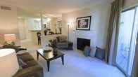 This single-floor residence offers an open-concept layout that connects the living area, dining space, and kitchen into one cohesive environment. The living room is anchored by a white-mantled fireplace, which serves as the room's focal point, flanked by decorative pillows and accented with framed wall art above. Plush wall-to-wall carpeting runs throughout the living area, contributing to a warm and comfortable ambiance. A dark wood coffee table and patterned armchair add contrast to the neutral tones of the space.

A partial wall with a structural column separates the living area from the kitchen, which is visible through a pass-through opening and features a raised breakfast bar with seating. The kitchen is fitted with light-toned cabinetry and appears to connect to an adjacent dining area. Ceiling fixtures provide consistent overhead lighting throughout the space. Large windows allow natural light to filter in, enhancing the open and airy feel of the interior. The overall design reflects a traditional aesthetic with neutral tones, creating a welcoming and functional living environment.