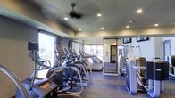 This fitness center is designed to offer a comprehensive workout experience in a modern, well-appointed setting. The space is outfitted with a robust selection of cardio equipment, including elliptical trainers, treadmills, and stair climbers, arranged in orderly rows to maximize usability and flow. A dedicated strength training area, equipped with Matrix-brand machines, is separated by a sleek glass-and-metal partition, providing a defined zone for resistance-based workouts. Large floor-to-ceiling windows line one wall, flooding the space with natural light and offering open views of the surrounding cityscape, creating an energizing atmosphere for workouts. The ceiling features recessed lighting complemented by a ceiling fan, ensuring consistent air circulation and a well-lit environment throughout. Flooring consists of durable, dark tile suited for high-traffic use. A wall-mounted television provides entertainment and visual engagement for cardio sessions. The overall ambiance is clean, contemporary, and purposefully functional, reflecting a facility designed to serve the needs of serious fitness enthusiasts. The thoughtful layout and quality equipment selection make this fitness center a standout amenity within the building.