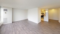 This single-floor unit presents a clean, contemporary design throughout its open layout. The living area is a standout feature, showcasing wide-plank gray wood-style flooring that extends seamlessly through the main living spaces. A row of large, black-framed windows lines the living area, flooding the space with natural light and offering expansive views of the surrounding urban cityscape.

The kitchen is well-appointed with a modern aesthetic, combining white upper cabinetry with warm wood-toned lower cabinets and light countertops. A full suite of stainless steel appliances includes a refrigerator, electric range, over-the-range microwave, and dishwasher. Light-toned tile flooring and a single-basin undermount sink with a contemporary faucet complete the functional workspace. A partial wall separates the kitchen from the living area, maintaining an open and connected feel.

The bathroom features a neutral, understated design with beige tile surround in the tub and shower combination, chrome fixtures, and clean white finishes throughout. The overall ambiance of the space is bright, modern, and move-in ready.