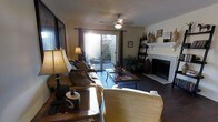 This single-floor residence offers a thoughtfully arranged layout with a warm and inviting atmosphere throughout. The entryway provides an immediate view into the living area, framed by a half-wall partition and accented by decorative artwork, setting a refined tone from the moment you arrive. Dark hardwood floors run throughout, adding richness and contrast to the neutral, light-toned walls.

The living room serves as the heart of the home, generously proportioned to accommodate comfortable seating arrangements centered around a wood-and-metal coffee table. A tile-surround fireplace anchors one wall, complemented by a decorative ladder shelf and tasteful accents. Natural light pours into the space through large sliding glass doors fitted with drape panels, offering direct access to a private outdoor patio area with greenery visible beyond. A ceiling fan overhead adds both function and a casual elegance to the room.

Built-in shelving is visible further into the home, hinting at additional storage and display opportunities. The textured ceiling adds architectural character, while recessed lighting and warm lamp fixtures contribute to a layered and cozy ambiance. Overall, this space combines practical design with comfortable, livable charm.