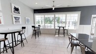 The Spring Creek clubhouse is a thoughtfully designed single-floor community gathering space that blends modern aesthetics with functional comfort. The main lounge area is anchored by expansive grid-pane windows that flood the room with natural light while offering direct views of the outdoor pool area and surrounding neighborhood — creating a seamless connection between indoor and outdoor living. Large-format light-toned tile flooring runs throughout, lending a clean, open feel to the space. High-top bistro tables paired with sleek black metal bar stools are arranged throughout the room, providing a casual yet stylish setting ideal for socializing or working. A ceiling fan with a brushed metal finish is centrally mounted, ensuring year-round comfort. The walls feature a striking two-tone design — crisp white wainscoting on the lower half contrasted by a bold dark accent band near the ceiling — adding a contemporary touch to the overall design. Thoughtful wall art, including a vintage-inspired typewriter print, contributes personality and warmth to the space. Recessed lighting complements the natural light, ensuring the room remains bright and welcoming at any time of day. Overall, the clubhouse offers a comfortable and inviting atmosphere for residents to gather and connect.