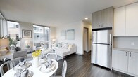 This single-floor space showcases a contemporary open-concept design that connects the living, dining, and kitchen areas into one cohesive, functional environment. Dark hardwood-style flooring runs throughout, providing a striking contrast against the crisp white walls and light-toned furnishings.

The living area is bathed in natural light from large floor-to-ceiling windows that offer views of the surrounding urban streetscape. A neutral-toned sectional sofa anchors the space, complemented by a clean white coffee table and soft decorative accents in blue and yellow.

Adjacent to the living area, a round white dining table with metal chairs creates an intimate setting for meals, with fresh floral arrangements adding a welcoming touch.

The kitchen is efficiently integrated into the open layout, featuring tall two-tone cabinetry in light wood and dark finishes that extend to the ceiling for maximum storage. A stainless steel refrigerator adds a modern, polished finish to the space.

A hallway visible in the background suggests additional private spaces beyond the main living area. Overall, the unit presents a clean, modern aesthetic with thoughtful use of light, contrast, and functional design.