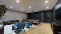 This single-floor amenity lounge is thoughtfully designed to serve as a social and recreational hub. The space is anchored by a solid wood-finished pool table positioned beneath a sleek, multi-arm pendant chandelier that provides focused task lighting while adding a contemporary aesthetic focal point. The flooring throughout is light-toned hardwood, lending warmth and a clean, modern feel to the room.

A seating area in the foreground features plush teal velvet armchairs arranged atop a patterned area rug, offering a comfortable gathering spot for conversation or relaxation. To the rear, bar-height tables with matching stools provide an additional casual seating option. A leather sofa is tucked against the wall, further expanding the lounge capacity.

The walls feature a striking contrast between light grey and a dark charcoal textured brick accent wall, contributing depth and visual interest to the space. Recessed lighting complements the pendant fixture, ensuring the room is well-lit throughout. A framed mirror on the far wall enhances the sense of openness. Overall, the space balances comfort and style in a well-appointed, multipurpose lounge environment.