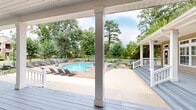 Located at 6650 Cottage Hill Rd in Mobile, Alabama, this single-story property offers an inviting blend of classic Southern architecture and thoughtfully designed outdoor amenities. The building features warm tan lap siding accented by crisp white trim, white columns, and a covered front porch with broad painted steps — creating a welcoming, traditional aesthetic. French doors with transom windows line the facade, allowing abundant natural light to flow between the interior and the expansive outdoor spaces.

The outdoor area is a standout feature of this property. A generously sized rectangular swimming pool takes center stage, surrounded by a handsome brick paver deck appointed with lounge chairs, decorative planters, and a small seating area — ideal for relaxation and outdoor gatherings. A white vinyl fence encloses the pool area, providing both safety and a clean visual boundary against the backdrop of towering mature trees that lend the space a private, retreat-like atmosphere.

Beyond the pool, a wooden pergola nestled among lush plantings offers a shaded secondary gathering spot. Manicured landscaping with established shrubs, ornamental grasses, and flowering plants lines the pathways and borders throughout the grounds. The combination of covered porch space, open concrete hardscape, and verdant greenery creates a seamless indoor-outdoor experience that captures the charm of the Alabama Gulf Coast lifestyle.