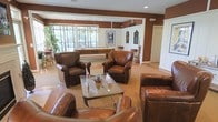 This single-floor recreational space is designed to offer both comfort and entertainment in a warm, inviting atmosphere. The lounge area is anchored by a generous seating arrangement of rich brown leather armchairs grouped around a glass-top coffee table, creating an ideal setting for relaxed conversation or casual gatherings. The deep brown accent walls contrast with white trim and architectural details, lending the space a classic, club-like aesthetic.

Toward the rear of the room, a full-size billiards table takes center stage beneath a hanging pendant light fixture, providing a dedicated games area that adds a social and recreational dimension to the space. Large floor-to-ceiling windows along the back wall flood the billiards zone with natural light and frame views of lush outdoor greenery, enhancing the overall sense of openness. A set of bar-height stools and a counter area nearby offer an additional spot for guests to gather. Framed artwork and decorative wall pieces are displayed throughout, adding personality and visual interest. The flooring transitions between hardwood-style surfaces and area rugs, defining the different functional zones while maintaining a cohesive, comfortable environment throughout.