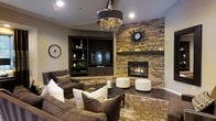 This single-story space showcases a thoughtfully designed interior that balances elegance with comfort. The main living area commands attention with its soaring ceilings, creating an expansive, airy atmosphere that draws the eye upward toward a statement chandelier featuring crystal and metallic accents. Dark hardwood flooring flows throughout, providing a rich contrast to the neutral warm-toned walls.

The living space is anchored by a striking stacked-stone accent wall housing a built-in gas fireplace, which serves as the room's focal point. Flanking the fireplace, a dark-finished built-in entertainment unit with open shelving provides both functional storage and display space. Recessed lighting complements the chandelier, ensuring well-distributed illumination throughout the area.

Furnishings include plush dark-brown upholstered sofas and accent chairs adorned with layered decorative pillows in neutral and patterned tones. A textured shag area rug and a round dark-finished coffee table complete the seating arrangement. White cylindrical ottomans near the fireplace add versatile functionality. Adjacent rooms are visible through open doorways, suggesting a well-connected and functional floor plan throughout the space.