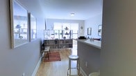 This single-floor space showcases a well-designed open-concept layout that effortlessly connects the kitchen and living areas. Light hardwood flooring runs throughout, lending warmth and a sense of continuity to the space. The kitchen features a raised bar counter with barstool seating, offering a casual dining option while maintaining a visual connection to the living area — ideal for both everyday living and entertaining.

The living area is anchored by a large, bright window that invites generous natural light, creating an airy and welcoming atmosphere. Soft, neutral gray walls complement the light tones of the floors, giving the space a clean and modern aesthetic. A distinctive open shelving unit serves as a stylish room divider, providing storage and display space while maintaining the open feel of the layout.

Additional decorative touches, including framed mirrors, wall art, and a patterned accent rug, add personality and depth to the space. The overall ambiance is light, contemporary, and thoughtfully arranged, making efficient use of the available space while maintaining a comfortable and inviting character throughout.