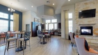 This single-floor clubhouse and fitness center is designed to serve as a premier amenity space, combining functionality with contemporary style. The main gathering area features dramatically high ceilings with a striking geometric soffit detail, creating a sense of grandeur and openness throughout. Light wood-tone flooring runs seamlessly across the space, lending a clean, modern aesthetic.

The fully equipped kitchen is a standout feature, outfitted with dark espresso cabinetry, stainless steel appliances, and ample counter space. Pendant lighting in teal accents adds a pop of color and visual interest above the kitchen island, complementing the overall design palette of blues, grays, and warm neutrals.

Natural light floods the interior through large, multi-pane windows dressed with patterned curtains, creating a bright and welcoming atmosphere. A textured stone or brick accent column near the window area introduces an element of architectural contrast and warmth.

The communal dining and lounge area is furnished with stylish bar-height seating, including mixed-material chairs with patterned cushions, ideal for socializing or casual gatherings. Recessed lighting throughout ensures the space remains well-lit at all hours. Overall, the clubhouse exudes a polished, resort-inspired ambiance tailored for community use.