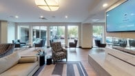 Located at 3900 Excelsior Blvd in St. Louis Park, Minnesota, this single-floor amenity lounge is designed with a contemporary aesthetic that balances comfort and functionality. The space is anchored by a striking marble-clad, double-sided gas fireplace with a wall-mounted television above, serving as a central architectural focal point that visually divides the room into distinct zones.

The lounge area features hardwood-style flooring throughout, recessed lighting, and a large geometric area rug anchoring a seating arrangement of stylish accent chairs and a low-profile coffee table. Floor-to-ceiling windows along one wall flood the space with natural light and offer views of the surrounding landscaping and exterior grounds, creating an open and airy atmosphere.

Adjacent to the lounge, a row of semi-private dining booths with dark wood panel walls, built-in bench seating, and individual televisions provides an intimate setting for smaller gatherings. A well-appointed wet bar and kitchenette area features dark cabinetry, white quartz countertops, a sink, beverage refrigerator, coffee station, hexagonal tile backsplash, and a large communal island with bar-height seating illuminated by bold brass pendant lights, making it ideal for entertaining and social gatherings.