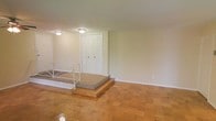This single-floor space offers a clean and functional layout designed for comfortable everyday living. The main living area is defined by warm parquet hardwood flooring in a classic block pattern, lending a timeless character to the interior. A raised entryway platform, finished with carpeted steps and framed by white metal railings, creates a subtle architectural transition between the entry and the main floor level. A set of traditional six-panel double doors serves as the front entry, providing a sturdy and classic focal point to the space. The walls throughout are finished in a soft neutral tone, creating a bright and airy atmosphere that is well-suited to a variety of décor styles. Flush-mount ceiling fixtures provide consistent overhead lighting across the space. Clean white baseboards and trim offer a crisp contrast to the warm flooring below. The open layout of the main area allows for flexible furniture arrangement, making it adaptable to both living and dining configurations. Overall, the space presents a straightforward and well-maintained interior with practical finishes and a welcoming, understated ambiance throughout its single level.