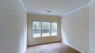 This single-floor space presents a clean, well-maintained interior defined by its warm, neutral palette and thoughtful architectural details. The room is finished with smooth hardwood-style flooring in a light wood tone, contributing to a bright and welcoming atmosphere throughout. Crown molding lines the perimeter where the walls meet the ceiling, adding a refined touch to the otherwise understated design.

A trio of double-hung windows positioned along one wall serves as a central feature of the space, drawing in generous amounts of natural light and providing pleasant views of surrounding greenery and trees. The windows are fitted with horizontal blinds, offering flexible light control while maintaining a tidy appearance.

The walls are painted in a soft, warm beige tone that complements the flooring and enhances the overall sense of openness. Crisp white baseboards and window trim provide clean contrast throughout the room. A ceiling-mounted HVAC vent ensures efficient climate control, and an electrical outlet is conveniently placed along the wall. The overall ambiance is light, airy, and move-in ready, offering a versatile blank canvas suitable for a variety of uses.