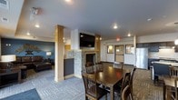 This single-floor communal space is thoughtfully designed to accommodate both casual socializing and relaxed downtime. At the heart of the room stands a striking stone-clad fireplace, which serves as a natural focal point and adds warmth and character to the overall ambiance. A wall-mounted flat-screen television is positioned above the fireplace, making it an ideal setting for group viewing or entertainment.

The space is divided into two distinct functional zones. A dining area features a solid dark wood table surrounded by matching wood-and-upholstered chairs, offering ample seating for meals or group gatherings. Adjacent to it, a comfortable lounge area is furnished with dark leather seating and accented by a warm table lamp, creating a more relaxed and intimate atmosphere.

Structural wood-trimmed columns add architectural interest and help define the zones within the open layout. The patterned carpeting throughout ties the design together with a modern touch. Recessed lighting provides consistent, even illumination across the space. A door with an emergency exit sign indicates accessibility and safety compliance. The overall design balances practicality with comfort, making this a versatile and inviting communal environment suited for a variety of uses.