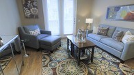 This single-story space showcases a thoughtfully arranged living area that combines comfort with a cohesive aesthetic. The room features warm hardwood flooring as its foundation, over which a large, eye-catching area rug with a bold floral and medallion pattern in blues, greens, and browns anchors the seating arrangement. Natural light floods the space through a set of full-length French doors fitted with white horizontal blinds, creating a bright and welcoming atmosphere. The living area is furnished with a powder-blue sofa accented by decorative patterned pillows, paired with a matching armchair and ottoman that together form an inviting conversation area. A dark wood coffee table with lower shelving serves as the central functional piece, styled with glass decanters and a mirrored tray. A coordinating end table supports a ceramic table lamp, adding warm ambient lighting. The walls are finished in a soft taupe tone, complemented by tasteful wall art. The overall ambiance is relaxed yet polished, with a neutral base palette elevated by carefully selected accent pieces that bring depth and visual interest to the room.