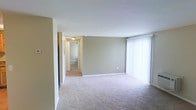 This single-story space is designed with a clean, functional layout that maximizes usability. The main living area is finished with wall-to-wall beige carpeting, providing a warm and comfortable foundation underfoot. Neutral greige-toned walls create a versatile backdrop, complementing the white trim and ceilings throughout. A large window dressed with vertical blinds dominates one wall, allowing for adjustable natural light and offering a bright, open atmosphere. The open layout of the living area provides ample space for a variety of furniture arrangements. A hallway visible from the main living area connects to additional rooms, suggesting a well-organized floor plan. The corridor is finished with the same neutral color palette and carpeting, maintaining visual consistency throughout the space. Ceiling-mounted lighting fixtures in the hallway ensure adequate illumination. The white baseboards and trim throughout the space provide a crisp contrast against the soft wall tones, adding a polished finish to the overall interior. The space presents a move-in ready, straightforward aesthetic suited for a range of uses.