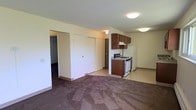This single-floor unit presents an open-concept layout that seamlessly connects the living area to the kitchen. The living space is finished with wall-to-wall carpet in a neutral tone, providing a comfortable underfoot feel, while the kitchen and surrounding areas transition to a smooth, easy-to-clean hard floor surface.

The kitchen is equipped with dark brown wood cabinetry, offering ample storage above the countertop. A white gas range and a full-sized white refrigerator are included, making the space move-in ready. An overhead range hood is mounted beneath the upper cabinets, providing ventilation during cooking. The kitchen layout is compact yet functional, with enough counter space for everyday meal preparation.

Adjacent to the living area, two closet doors are visible along a shared wall, offering generous storage options. A doorway leads to an additional room or hallway beyond, adding to the overall utility of the unit. Flush-mount ceiling light fixtures illuminate the space throughout. The walls are finished in a clean, neutral white, contributing to a bright and simple interior aesthetic.