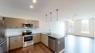 This single-floor space showcases a contemporary design with a thoughtful open-concept layout. The kitchen serves as the centerpiece, featuring a full suite of stainless steel appliances including a french door refrigerator, range with microwave above, and dishwasher integrated into the peninsula island. Taupe flat-front cabinetry extends from floor to ceiling, complemented by a white subway tile backsplash and dark countertops that carry through to the island. Three elegant Edison-style pendant lights hang above the peninsula, adding a refined touch. Recessed lighting and hardwood-style flooring complete the kitchen and living areas, which flow seamlessly together. A large window in the living area invites natural light and outdoor views. The bedrooms feature plush carpeting and ceiling fans, with one room benefiting from a window fitted with a roller shade. The bathroom is well-appointed with a dark-topped vanity, undermount sink, pendant lighting over a large mirror, and a full soaking tub with shower. The clean white walls and neutral palette throughout create a bright, modern ambiance.