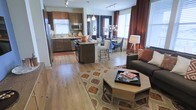 This single-floor residence showcases an open-concept layout that integrates the kitchen, dining, and living areas into one cohesive, flowing space. The kitchen features rich dark-wood cabinetry, stainless steel appliances including a refrigerator and microwave, a mosaic tile backsplash, and a central island with a granite countertop and bar seating — ideal for casual dining or entertaining. Pendant lighting hangs above the island, complementing the track lighting that runs throughout the space. The dining area is positioned near large windows dressed in bold rust-colored drapes, allowing generous natural light to fill the room. A round dining table surrounded by upholstered chairs creates an inviting setting. The living area transitions naturally from the dining space, featuring wood-look flooring, a patterned area rug, an octagonal coffee table, and a dark upholstered sofa accented with warm-toned throw pillows. The combination of warm wood finishes, curated furnishings, and an abundance of natural light gives the space a welcoming and well-appointed ambiance throughout.