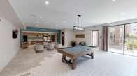 This thoughtfully designed single-floor amenity space in Houston, TX (77055) offers a contemporary and inviting atmosphere well-suited for socializing and recreation. The layout flows seamlessly between distinct activity zones, all unified by large white hexagonal tile flooring that lends a clean, modern aesthetic throughout.

The game area serves as a central focal point, anchored by a rustic wood-framed pool table beneath an industrial-style rectangular pendant light. Dual wall-mounted flat-screen televisions are positioned nearby, supported by recessed lighting across the ceiling, creating an ideal setting for entertainment.

Adjacent to the game area is a well-appointed lounge zone featuring upholstered armchairs and café-style seating arrangements. A bold teal accent wall adds a vibrant pop of color, defining the kitchen area, which is equipped with light wood cabinetry featuring gold hardware, a stainless steel refrigerator, a dishwasher, and a large island with a white stone countertop. Black pendant lights hang above the island, complemented by wire barstools in a warm metallic finish.

Near the entry, a sitting area with deep navy accent chairs, a textured wall art piece, and a glazed door with outdoor views creates a welcoming transition into the space. The overall ambiance is modern, functional, and polished.