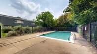 This outdoor pool area offers a well-maintained recreational space designed for comfortable outdoor use. The centerpiece is a large rectangular swimming pool with clear turquoise water, featuring marked depth indicators along its edges and metal handrail entries for safe access. A life ring is mounted poolside, reflecting standard safety provisions.

The pool is enclosed by a sturdy wrought iron fence that defines the perimeter while maintaining an open, airy feel. The surrounding concrete deck provides ample space for lounging and movement, and several metal tables and chairs are positioned along the edges, offering shaded seating beneath the canopy of mature deciduous trees that line the boundary. These trees contribute significantly to the ambiance, providing natural shade and a lush, green backdrop that softens the hardscape.

A compact brick utility building with a red door is situated at one end of the pool area, likely housing mechanical equipment or changing facilities. The overall setting is bordered by adjacent low-rise brick buildings, suggesting a shared community amenity. The combination of practical safety features, generous deck space, and verdant tree coverage creates a functional and inviting outdoor environment well-suited for recreation and relaxation.
