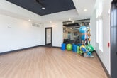 Located at 5039 W Broad St in Sugar Hill, GA, this single-floor fitness studio offers a well-designed, open-concept workout environment built for versatile exercise and group classes. The space is defined by warm hardwood-style flooring that spans the entire room, creating a clean and functional foundation for a wide range of activities.

One of the studio's most striking features is its expansive wall of floor-to-ceiling mirrors, which visually amplifies the space and provides exercisers with full-body form feedback. A row of spin bikes is neatly arranged along the mirrored wall, accompanied by a mounted flat-screen display for guided workouts or media. The high ceilings are finished in a bold navy blue, creating a strong contrast against the crisp white walls and adding a modern, energetic aesthetic to the interior.

Large multi-pane windows along the exterior wall allow abundant natural light to pour into the studio, offering views of the surrounding outdoor environment and a bright, uplifting atmosphere. Additional equipment and accessories — including medicine balls, stability balls, and resistance tools — are organized throughout the room, supporting diverse training modalities. Recessed lighting supplements the natural light, ensuring the space is well-illuminated at all times. A secondary door leads to an adjacent fitness area, making this studio part of a broader wellness facility.