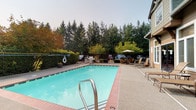 This well-appointed outdoor recreational area centers around a generously sized swimming pool with clear turquoise water, framed by a brick-accented surround and a textured concrete deck. The pool is easily accessible via stainless steel handrails and entry steps, making it welcoming for all users. Adjacent to the pool, a separate hot tub offers a more intimate relaxation option, also finished with brick tile edging and equipped with its own handrail for safe entry.

The surrounding deck provides ample space for lounging, with multiple sling-style chaise loungers positioned to take in the open-air setting. A shaded seating area with a patio umbrella, additional chairs, and a BBQ grill creates an inviting space for outdoor gatherings. The entire area is enclosed by a black iron fence, lending both security and a clean aesthetic.

The backdrop is defined by a dense stand of tall evergreen and deciduous trees, providing natural privacy and a serene, Pacific Northwest atmosphere. The building exterior, clad in warm taupe lap siding with white-trimmed windows, houses practical amenities including a wall-mounted drinking fountain and clearly marked restroom facilities with separate-entry doors. Posted signage with pool rules ensures the space remains organized and well-managed for all users.