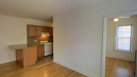 This single-floor space offers a practical and straightforward layout, with a living area that flows openly into an adjacent kitchen. The living area features warm-toned hardwood flooring and clean, neutral white walls, creating a bright and simple backdrop that is easy to furnish and personalize. White trim and baseboards provide a crisp contrast throughout.

The kitchen is accessible directly from the main living space and is appointed with medium-toned wooden cabinetry that provides ample storage. A granite or stone-look countertop adds a touch of durability and texture, complemented by a matching tile backsplash that ties the aesthetic together. A white gas range sits beneath an overhead exhaust hood, offering functional cooking capacity. The kitchen floor transitions to tile, clearly delineating the two spaces while maintaining a cohesive overall feel.

A doorway with white trim visible along the right side of the space suggests access to additional rooms beyond the main area. The overall ambiance is clean, functional, and move-in ready, with warm material choices lending a comfortable and lived-in character to the space.