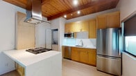 This single-floor residence showcases a thoughtful blend of warm natural materials and contemporary finishes. The kitchen serves as a standout centerpiece, featuring rich wood cabinetry, a quartz waterfall island with a gas cooktop, a sleek stainless steel hood, and a full suite of stainless steel appliances including a refrigerator, dishwasher, and built-in microwave. A marble-style backsplash and under-cabinet lighting add a refined touch. A stacked washer and dryer unit is conveniently tucked nearby. Distinctive wood-planked ceilings with exposed beams run throughout portions of the home, lending warmth and architectural character to the open living areas. Light hardwood flooring flows seamlessly across all rooms, enhancing the bright, airy atmosphere. The bedrooms are clean and neutral, with white walls and ample natural light; one features a frosted-glass sliding closet door, while others benefit from the dramatic beamed ceilings shared with the main living space. A window in one bedroom allows natural light to filter in. The bathroom is finished with large-format beige tile, a decorative mosaic border, and a glass-enclosed soaking tub and shower combination, offering a serene, spa-like retreat.