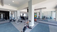 This fitness center offers a spacious, open layout designed to accommodate a wide variety of workout routines. The space features a clean, contemporary aesthetic with white walls, structural columns, and a combination of carpet and rubberized flooring that defines distinct workout zones. Cycling bikes and cardio machines are positioned toward the front of the space, providing easy access for warm-up routines. Further into the room, a well-stocked free weight area includes a tiered dumbbell rack and colorful bumper plate storage, catering to strength training needs. A wall of mirrors lines one section of the space, enhancing the sense of openness and allowing users to monitor their form during exercises. A flat-screen television is mounted near the ceiling, offering entertainment during workouts. Recessed lighting throughout maintains a bright and energizing atmosphere. The thoughtful arrangement of equipment ensures ample circulation space between stations, making the facility feel both functional and comfortable for simultaneous use by multiple individuals.