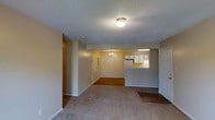 This single-floor residence offers a straightforward and functional layout designed for comfortable everyday living. The main living area is spacious and open, featuring neutral-toned carpet flooring and textured ceilings that lend a classic residential feel. A flush-mount ceiling light fixture illuminates the living space, while a small chandelier adds a decorative touch in the adjacent dining area, which transitions to hard flooring. The open floor plan allows the living, dining, and kitchen areas to flow seamlessly into one another. The kitchen is accessible via a pass-through counter, providing a convenient connection between the cooking and dining spaces. White cabinetry lines the kitchen walls, complemented by a refrigerator and overhead fluorescent lighting that keeps the area bright and practical. Neutral beige walls throughout the unit create a clean, cohesive backdrop that is easy to personalize. A front entryway with a paneled door provides a welcoming point of entry, while additional doorways lead to other areas of the unit. The overall ambiance is understated and functional, making it a practical space well-suited for a range of living arrangements.