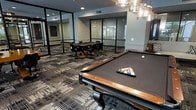 This single-floor game room is designed as a dedicated recreational space, combining modern architectural elements with comfortable amenities for social gatherings. The centerpiece of the room is a full-sized pool table with a rich wood finish, dark felt surface, and ornate carved legs, positioned beneath a stylish overhead light fixture. Racked and ready for play, the table sets an inviting tone for the space.

Adjacent to the billiards area, a card or poker table with matching dark chairs provides a secondary gaming option, ideal for small group gatherings. The flooring throughout features a patterned carpet in neutral gray and charcoal tones, adding texture and visual interest underfoot.

A defining architectural feature is the floor-to-ceiling glass partition wall framed in black metal, which encloses the space while maintaining a sense of openness and allowing natural light to filter in from adjacent areas. Recessed ceiling lighting supplements the ambient illumination. The overall aesthetic is contemporary and polished, creating a well-appointed environment suited for leisure and entertainment.