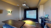 This thoughtfully designed single-floor amenity space offers a diverse range of features tailored for both recreation and productivity. The standout highlight is a dedicated golf simulator room, equipped with a ceiling-mounted projector, a large projection screen framed by black curtains, and a synthetic turf hitting mat — all set against warm neutral walls and rich wood-look flooring, creating an immersive entertainment experience.

Adjacent to the simulator, a well-appointed business center provides workstations with desktop computers, a wall-mounted television displaying branded content, and a coffee station, making it a convenient hub for guests or residents needing to stay connected. The area benefits from ample natural light streaming through large windows fitted with horizontal blinds, offering views of the surrounding landscape.

The open lounge and dining area extends seamlessly toward a glass-enclosed patio, flooding the interior with additional light and providing easy indoor-outdoor access. Warm golden accent walls contrast with the drop-panel ceilings and dark hardwood-style flooring throughout.

A fully equipped kitchen supports the communal space with a full-size refrigerator, electric range, microwave, stainless steel range hood, and a double-basin sink with generous counter space and cabinetry — ideal for hosting events or everyday use.