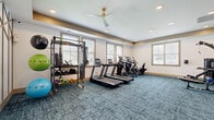 This fitness center offers a spacious, single-level layout designed to accommodate a comprehensive workout experience. The main exercise area is finished with durable, teal-toned carpet tiles that lend the space a clean, modern aesthetic. Recessed lighting and ceiling fans with gold accents provide both ample illumination and effective air circulation throughout the room.

Multiple windows with blinds line the walls, allowing natural light to fill the space while maintaining a bright and energizing atmosphere. A distinctive geometric wall treatment featuring a repeating triangular pattern adds a stylish architectural detail that elevates the overall design.

The fitness floor is thoughtfully equipped with a variety of cardio machines, including treadmills and elliptical trainers, as well as a multi-station cable and weight machine for strength training. A dedicated free weights area offers kettlebells, dumbbells, and a SmartFit functional training rack stocked with resistance bands, medicine balls, and accessories. An adjustable workout bench and a rowing machine round out the strength and conditioning options.

A wall-mounted flat-screen television is positioned in a secondary area divided by glass partitions, creating a defined zone ideal for stretching or focused training. The space is accessible via glass-paneled entry doors and includes safety features such as clearly marked exit signage, offering a safe and well-organized environment for all fitness levels.