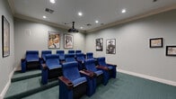 Located at 1900 Westview Blvd in Conroe, Texas, this dedicated home theater room delivers an authentic cinematic experience within a compact, well-appointed space. The room is designed with purpose and attention to detail, featuring rows of plush, blue upholstered theater-style seats complete with built-in cup holders and rich wood armrests — mirroring the comfort of a commercial movie house. Arranged in a tiered layout, the seating ensures unobstructed sightlines to the large projection screen mounted at the front of the room. A ceiling-mounted projector provides the visual centerpiece of the setup, while recessed lighting throughout the ceiling offers adjustable ambient illumination suited to any screening. The walls are adorned with framed classic Hollywood movie posters — including iconic titles such as Citizen Kane, Casablanca, and Rear Window — lending the room a nostalgic, cinephile character. Soft teal carpeting covers the floor, adding warmth and acoustic dampening. A wooden media cabinet positioned near the screen offers storage for equipment and accessories. Crown molding along the ceiling adds a refined architectural touch, and the neutral gray walls keep the focus on the screen. Two entry doors provide convenient access to adjoining spaces.