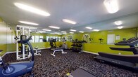 This fitness center is a single-floor facility designed to support a wide range of workout routines. The space is immediately distinguished by its vibrant chartreuse green walls, which lend a high-energy atmosphere throughout. A full-length wall mirror spans a significant portion of the room, visually expanding the space and allowing users to monitor their form during exercise.

The cardio section features two treadmills positioned along a windowed wall, where multiple windows with white blinds allow natural light to filter in and offer views of the surrounding greenery outdoors. An elliptical trainer, upright stationary bike, and additional cardio equipment round out the aerobic options.

For strength training, the center includes a multi-station cable machine, a weight bench, and a well-stocked dumbbell rack with a range of free weights. The equipment is thoughtfully arranged to maximize floor space and allow comfortable movement between stations.

The flooring throughout consists of a durable, patterned carpet with dark tones, well-suited to a high-traffic fitness environment. Overhead, flush-mounted oblong light fixtures provide consistent, bright illumination across the entire space. The facility also includes a dedicated entry door, ventilation grilles, and informational signage, reflecting a functional and user-focused design.