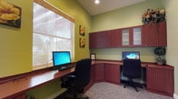 This single-floor business center is thoughtfully designed to provide a productive and comfortable workspace. The room features a built-in L-shaped desk finished in rich mahogany-toned wood, offering ample surface area for dual workstations, each equipped with a desktop computer and ergonomic office chair. Overhead, matching wall-mounted cabinetry with frosted glass and open shelving panels provide generous storage while maintaining a clean, organized aesthetic.

The walls are painted in a warm sage green, lending the space a calm and focused ambiance. A large window dressed with natural bamboo roller blinds allows soft, diffused natural light to fill the room, enhancing the welcoming atmosphere without harsh glare on screens. Recessed ceiling lighting supplements the natural light, ensuring consistent illumination throughout.

Decorative floral artwork adorns the walls, adding a subtle touch of warmth and personality to the otherwise professional setting. The floor is covered in neutral, low-pile carpeting that complements the overall color palette. The space is efficiently arranged to maximize functionality within its footprint, making it an ideal setting for focused work, research, or business tasks.