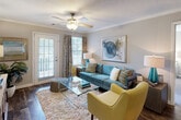 This single-floor residence offers a thoughtfully designed layout that balances comfort and functionality throughout. Dark hardwood-style flooring flows through the main living areas, creating a warm and cohesive aesthetic. The open-concept living room is bright and welcoming, featuring crown molding, a ceiling fan with light fixture, and a glass-paned door that opens to a private outdoor balcony, filling the space with natural light. The kitchen is well-equipped with light-toned cabinetry, stone-finish countertops, black appliances including a range and refrigerator, and a convenient breakfast bar with a sink that overlooks the main living area. The primary bedroom offers plush carpeting, generous natural light from large windows, and direct access to a bathroom featuring a wide vanity with a stone-finish countertop and large mirror. A second bedroom is configured for multiple occupants, complete with bunk beds and carpeted flooring, and is served by an adjacent bathroom with a single-sink vanity. Additional hallway space provides connectivity between rooms, accented by framed artwork and track lighting. Textured ceilings are present throughout, and the overall palette of soft grays and warm neutrals lends a calm, inviting ambiance to the entire residence.