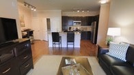 This single-floor residence offers a thoughtfully designed open-concept layout that connects the living, dining, and kitchen areas into one cohesive and functional space. The kitchen is a standout feature, outfitted with rich dark cabinetry, granite countertops, and a full suite of stainless steel appliances including a refrigerator, range, and microwave. A peninsula-style breakfast bar with seating for two provides a casual dining option while keeping the kitchen visually connected to the rest of the living space. Warm hardwood flooring flows throughout the main area, adding a sense of continuity and natural warmth. The living area is comfortably proportioned, anchored by a glass-top coffee table and area rug that define the space. Track lighting fixtures illuminate both the kitchen and living areas with clean, modern overhead lighting. Crown molding along the ceilings adds a refined architectural touch. Interior doors with white paneling lead to additional rooms, contributing to a clean and classic aesthetic. The overall ambiance is modern yet inviting, with a neutral color palette and quality finishes that create a polished, comfortable environment throughout.