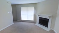 This single-floor residential space offers a clean, understated layout designed for comfortable everyday living. The living area serves as the focal point of the home, showcasing a classic white-painted brick fireplace with a simple mantel that adds warmth and character to the room. A large window fitted with horizontal blinds allows generous amounts of natural light to pour in, brightening the neutral, beige-toned walls and complementing the plush wall-to-wall carpeting underfoot. The soft carpet and muted color palette create a calm, neutral ambiance throughout, providing a versatile backdrop that suits a variety of interior styles. The room's smooth, continuous ceiling and clean baseboards reflect a well-maintained interior. The open feel of the living space, combined with the fireplace as a central architectural feature, gives the home a welcoming and functional atmosphere. Overall, this space presents a straightforward and practical single-floor layout with timeless design elements that offer both comfort and livability.