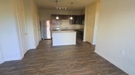 This single-floor space showcases a clean, modern design centered around an open-concept layout that seamlessly connects the kitchen and living areas. The dark hardwood-style flooring runs throughout, providing a warm and cohesive foundation that contrasts beautifully with the neutral-toned walls and white baseboards.

The kitchen is a standout feature, equipped with rich dark wood cabinetry that extends to the upper walls, offering generous storage. A central island with a light-colored countertop serves as both a functional workspace and a natural gathering point. Pendant lights hang above the island, complementing the track lighting overhead and creating a layered, well-lit environment. The kitchen also features stainless steel appliances, including a gas range, built-in microwave, and a full-size refrigerator, as well as a sink positioned beneath a window at the back counter.

The open living area flows naturally from the kitchen, offering a spacious and versatile footprint. The layout is practical and inviting, with clear sightlines throughout the main living space. Doorways visible from the main area suggest additional rooms, contributing to the overall functional design of this well-appointed single-floor residence.