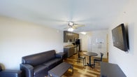 This single-floor space offers a practical and efficient open-plan layout that seamlessly combines a living area, kitchenette, and dining zone. The living area is anchored by a dark leather sofa paired with a rectangular coffee table, sitting atop warm hardwood floors that run throughout the space, lending a cohesive and inviting feel. A ceiling fan with an integrated light fixture is centrally positioned overhead, providing both air circulation and ambient lighting to the main living area. The kitchenette is neatly tucked toward the rear and features dark wood cabinetry, a stainless steel sink, a microwave, and full-size refrigerator and range appliances, offering all essential cooking and storage amenities. Adjacent to the kitchen, a compact round dining table with matching chairs provides a dedicated space for meals. A whiteboard mounted near the entryway adds a functional, practical touch. The space benefits from smooth, light-toned walls that reflect brightness and create an open, airy atmosphere. Overall, the layout is thoughtfully designed to maximize utility within a streamlined footprint, making it well-suited for everyday living.