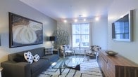 This thoughtfully designed single-floor space showcases a contemporary aesthetic with a keen eye for detail. The living area serves as the centerpiece, anchored by a sleek gray sofa adorned with patterned throw pillows and a striking glass-topped coffee table that adds an airy, modern touch. Track lighting runs across the ceiling, providing adjustable, layered illumination throughout the room. A large window floods the interior with natural light, framing a view of the surrounding building exterior and creating a bright, welcoming atmosphere. Two accent chairs in a bold black-and-white pattern flank a minimalist round side table near the window, offering an inviting conversational arrangement. A plush, textured area rug in a neutral tone grounds the seating area and adds warmth underfoot. The walls are adorned with curated artwork, including a large abstract canvas, while a sculptural metal wall accent introduces an artistic focal point. A warm-toned wooden credenza completes the space, providing functional storage with an elegant finish. The overall ambiance is chic and cohesive, blending modern furnishings with thoughtful decorative elements for a polished, livable interior.