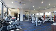 This fitness center offers a comprehensive workout environment designed to accommodate a variety of exercise needs. The space is finished with striking blue-striped carpet tiles that add a modern, energetic aesthetic to the room. Recessed lighting throughout the ceiling, complemented by a ceiling fan, ensures the area remains well-lit and comfortable during workouts. Large mirrors line the walls, amplifying the sense of space and allowing users to monitor their form. Natural light filters in through a row of windows along one wall, contributing to a bright and motivating atmosphere. The facility is stocked with a diverse selection of equipment, including elliptical machines and other cardio units, as well as a multi-station cable and strength training machine and a dedicated free weights area complete with a dumbbell rack. A water station is conveniently positioned within the space for easy access during workouts. The fitness center is accessed through a glass-paneled door, providing a clean, open visual connection to adjacent areas. White walls and a neutral ceiling keep the design clean and focused, while the overall layout allows for ample circulation between equipment stations.