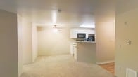 This single-floor space offers an open-concept design that seamlessly connects the living area to the kitchen. The living area features wall-to-wall carpet in a neutral beige tone, creating a warm and cohesive atmosphere throughout. A ceiling fan with an integrated light fixture is centrally positioned, providing both airflow and ambient lighting to the space.

The kitchen is accessible via a peninsula-style counter that serves as a natural divider between the two areas. White cabinetry lines the kitchen walls, offering ample storage, while black appliances—including a microwave and refrigerator—provide a subtle contrast. The kitchen benefits from recessed overhead lighting, ensuring a well-lit workspace. The transition from carpet to hard flooring marks the boundary between the living and kitchen areas.

The neutral color palette of beige and white walls throughout contributes to a bright, airy feel. The open layout and unobstructed sightlines between the kitchen and living area make the space feel connected and functional, well-suited for everyday living and casual entertaining.