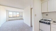 Located at 330 Oak Grove St in Minneapolis, MN, this efficiently designed unit offers a well-organized layout comprising a living room, kitchen, bathroom, and closet. The living area is generously proportioned for the overall footprint, featuring neutral carpet flooring, bright white walls, and large windows that invite abundant natural light while offering views of the surrounding neighborhood. Baseboard heating runs along the exterior walls, and a wall-mounted air conditioning unit ensures year-round comfort. The kitchen is thoughtfully appointed with white cabinetry extending to the ceiling for maximum storage, a gas range, single-basin sink, and a full-size refrigerator. Warm hardwood-style flooring in the kitchen and entryway adds a welcoming contrast to the carpeted living space. The bathroom is finished with classic white tile walls, vinyl flooring, a standard tub, and a window that provides natural ventilation. A dedicated closet offers practical storage. The clean, minimalist aesthetic throughout — defined by white walls and simple finishes — creates a bright, open ambiance that makes the space feel larger than its compact footprint suggests.