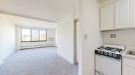 Located at 330 Oak Grove St in Minneapolis, MN, this efficiently designed unit offers a well-organized layout comprising a living room, kitchen, bathroom, and closet. The living area is generously proportioned for the overall footprint, featuring neutral carpet flooring, bright white walls, and large windows that invite abundant natural light while offering views of the surrounding neighborhood. Baseboard heating runs along the exterior walls, and a wall-mounted air conditioning unit ensures year-round comfort. The kitchen is thoughtfully appointed with white cabinetry extending to the ceiling for maximum storage, a gas range, single-basin sink, and a full-size refrigerator. Warm hardwood-style flooring in the kitchen and entryway adds a welcoming contrast to the carpeted living space. The bathroom is finished with classic white tile walls, vinyl flooring, a standard tub, and a window that provides natural ventilation. A dedicated closet offers practical storage. The clean, minimalist aesthetic throughout — defined by white walls and simple finishes — creates a bright, open ambiance that makes the space feel larger than its compact footprint suggests.