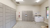 Located at 401 N Old Orchard Ln in Lewisville, Texas, this shared laundry facility offers a clean, functional, and well-organized space designed for everyday convenience. The room features durable speckled epoxy flooring throughout, bright white walls, and overhead fluorescent lighting that ensures the space remains well-lit at all times. Large windows allow natural light to filter in, offering views of the surrounding landscaped exterior. A dedicated laundry alcove houses commercial-grade top-load washers, equipped with digital controls for ease of use. Informational signage is posted on the walls to assist users with machine operation and facility guidelines. A standout feature of this space is its extensive package locker system, with multiple banks of gray steel lockers lining the walls — providing secure, convenient package storage and retrieval. An electronic locker kiosk with a built-in touchscreen interface allows for seamless access. Additional storage cabinetry and a seating area with a folding chair offer added practicality. A fire safety station is also present, reflecting attention to building safety standards. Glass-panel doors provide easy access to the outdoor surroundings, reinforcing a connected and accessible feel throughout this well-appointed communal facility.
