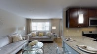 This single-floor residence showcases an open-concept layout that connects the living area and kitchen in a cohesive, functional design. The living room centers around a plush, light gray sofa accented with decorative pillows in neutral and yellow tones, complemented by a glass-topped coffee table with an ornate metal base that adds a refined touch. A patterned area rug in soft gray tones anchors the seating arrangement atop warm hardwood flooring that runs throughout the space. Large windows dressed with sheer, floor-length curtains allow generous natural light to fill the room, creating an airy and welcoming ambiance. The kitchen, visible from the living area, features rich dark wood cabinetry, granite countertops, and a light tile backsplash, offering a striking contrast to the softer tones of the living space. Sleek bar seating along the kitchen counter provides a casual dining option. The overall color palette—warm beiges, grays, and deep espresso tones—creates a sophisticated yet comfortable atmosphere well-suited for both everyday living and entertaining.