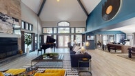 Located at 455 Arbor Pl in Nashville, TN, this single-floor community space delivers a sophisticated, music-inspired aesthetic that nods to its vibrant city roots. The main gathering area is anchored by dramatic vaulted ceilings with exposed dark beams, bold teal accent walls, and light wood-look flooring that flows throughout. A grand piano commands attention near a floor-to-ceiling stone fireplace, creating an impressive focal point framed by towering windows that draw in generous natural light. Eclectic décor — including a vinyl record wall accent, globe pendant lamps, and curated furnishings in navy, mustard, and gold — establishes a warm yet contemporary atmosphere. Multiple seating arrangements encourage socializing, while a dedicated dining area with sleek chairs and bistro-style tables offers a more intimate setting. A sunlit flex room with wraparound windows, carpeted floors, and ceiling fan provides a comfortable workspace or lounge area with views of the surrounding greenery. A digital kiosk near the entrance adds a modern, tech-forward convenience. With an abundance of glass doors opening to the outdoors and thoughtfully layered design details throughout, this space strikes an ideal balance between community functionality and upscale Nashville style.